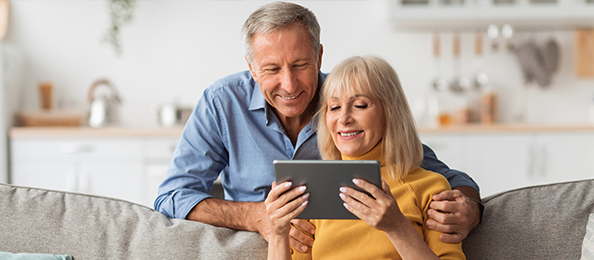 An older man and woman smile at a tablet while lounging on a couch.